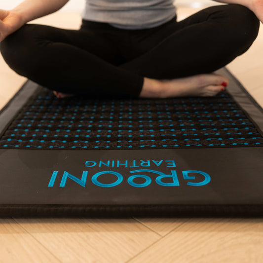 Person sitting cross-legged on a black Grooni PEMF mat with blue dots, meditating in a bright room with wooden flooring.