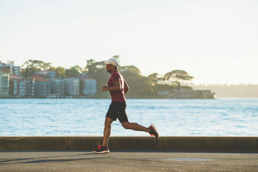 Man jogging along a sunny waterfront path, wearing athletic gear and a white cap.