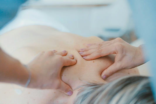Close-up image of a person receiving a therapeutic back massage. A massage therapist's hands are applying pressure to the upper back and shoulder area, suggesting relaxation, muscle tension relief, and wellness benefits.