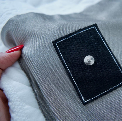 Grounding blanket with a black square patch featuring a silver button held by a hand with red nail polish