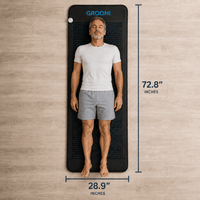 A man lies flat on a long, narrow black mat with measurement lines and text showing dimensions: 72.8 inches tall and 28.