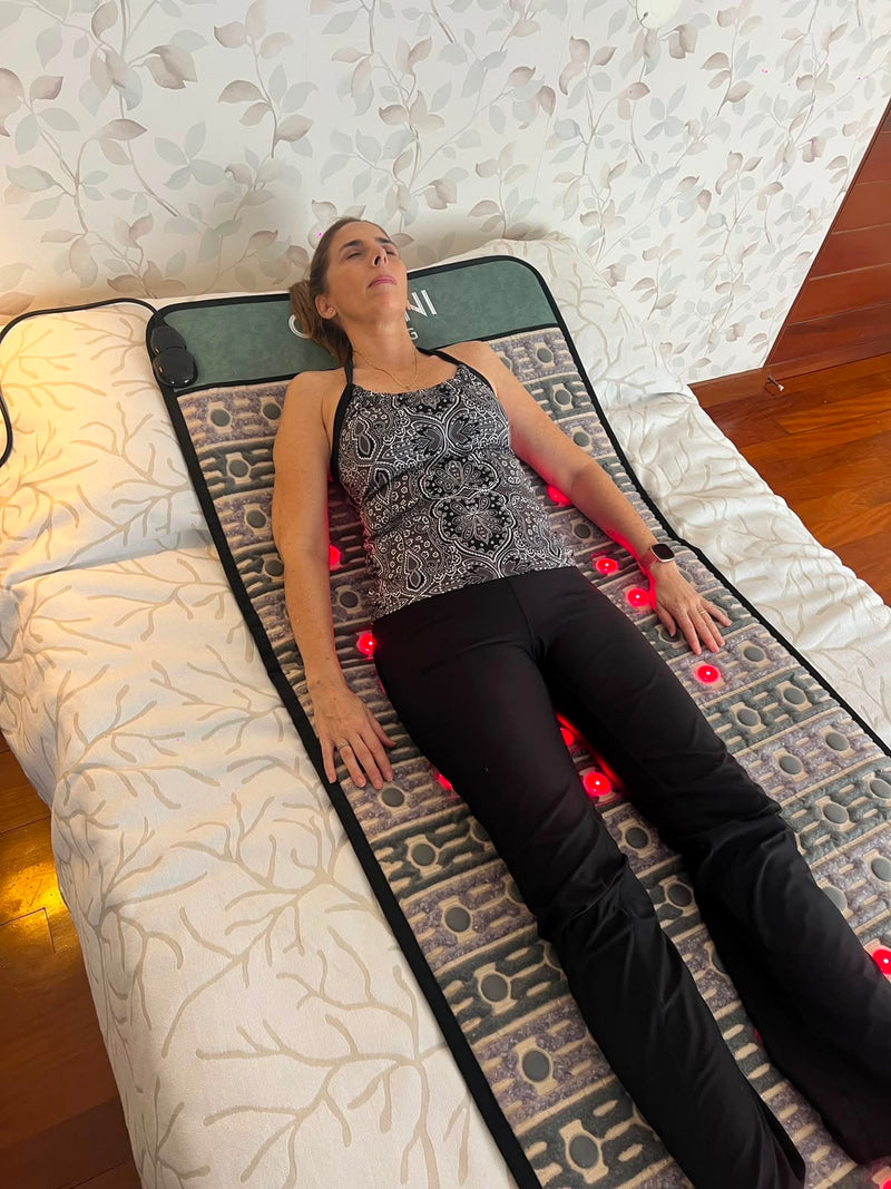 A woman lies on a patterned massage or heat therapy mat with visible red light dots, positioned on a bed in a home setting.