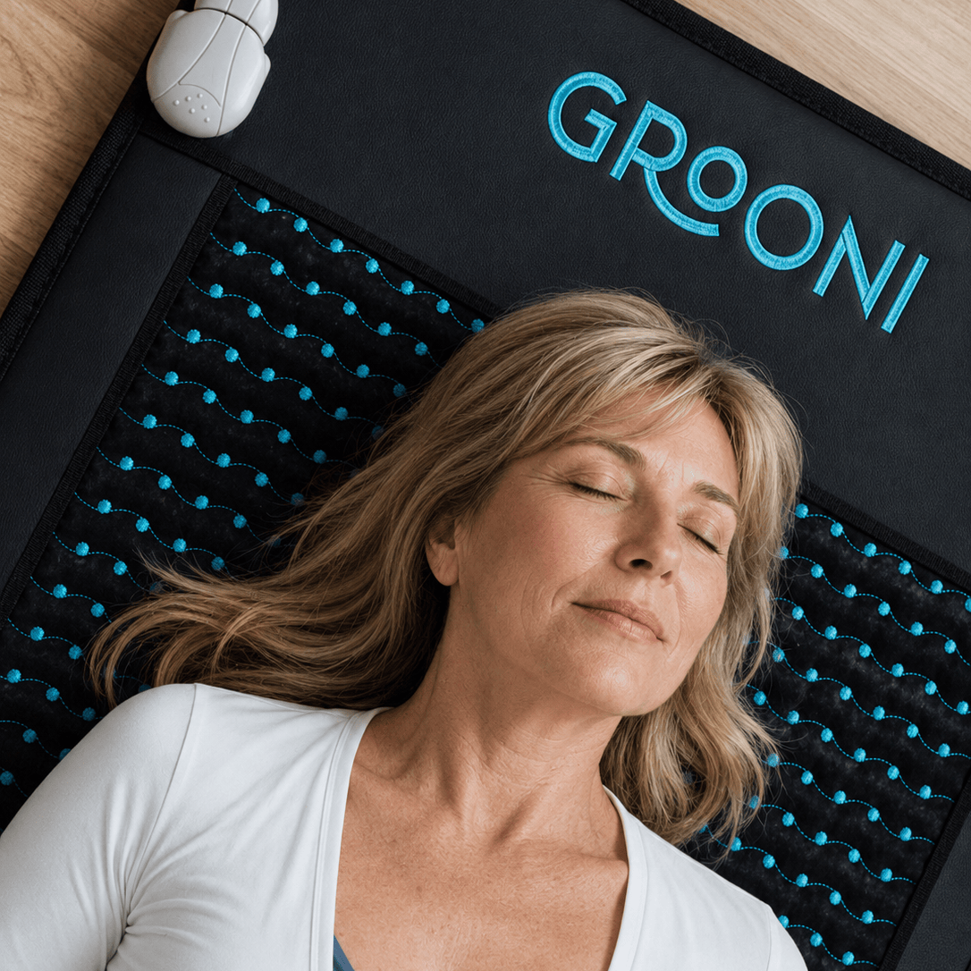 A woman with closed eyes rests on a black GROONI massage cushion with blue-accented nubs, shown in a home-like setting with a wooden floor visible.