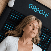 A woman with closed eyes rests on a black GROONI massage cushion with blue-accented nubs, shown in a home-like setting with a wooden floor visible.